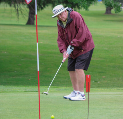 Man wearing a bucket hat and windbreaker jacket putting on the green toward the hole with the flag in it.